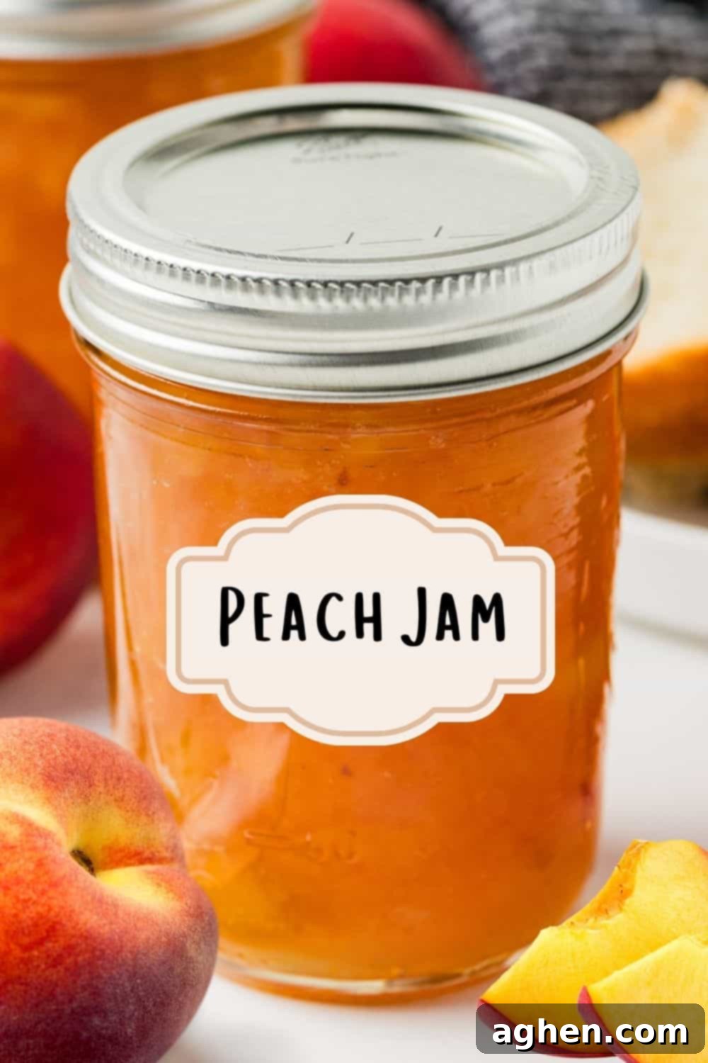A sealed mason jar labeled “Peach Jam” sits on a marble surface surrounded by fresh peaches, a slice of bread, and another jar in the background. Freshly sealed jar of homemade peach jam.