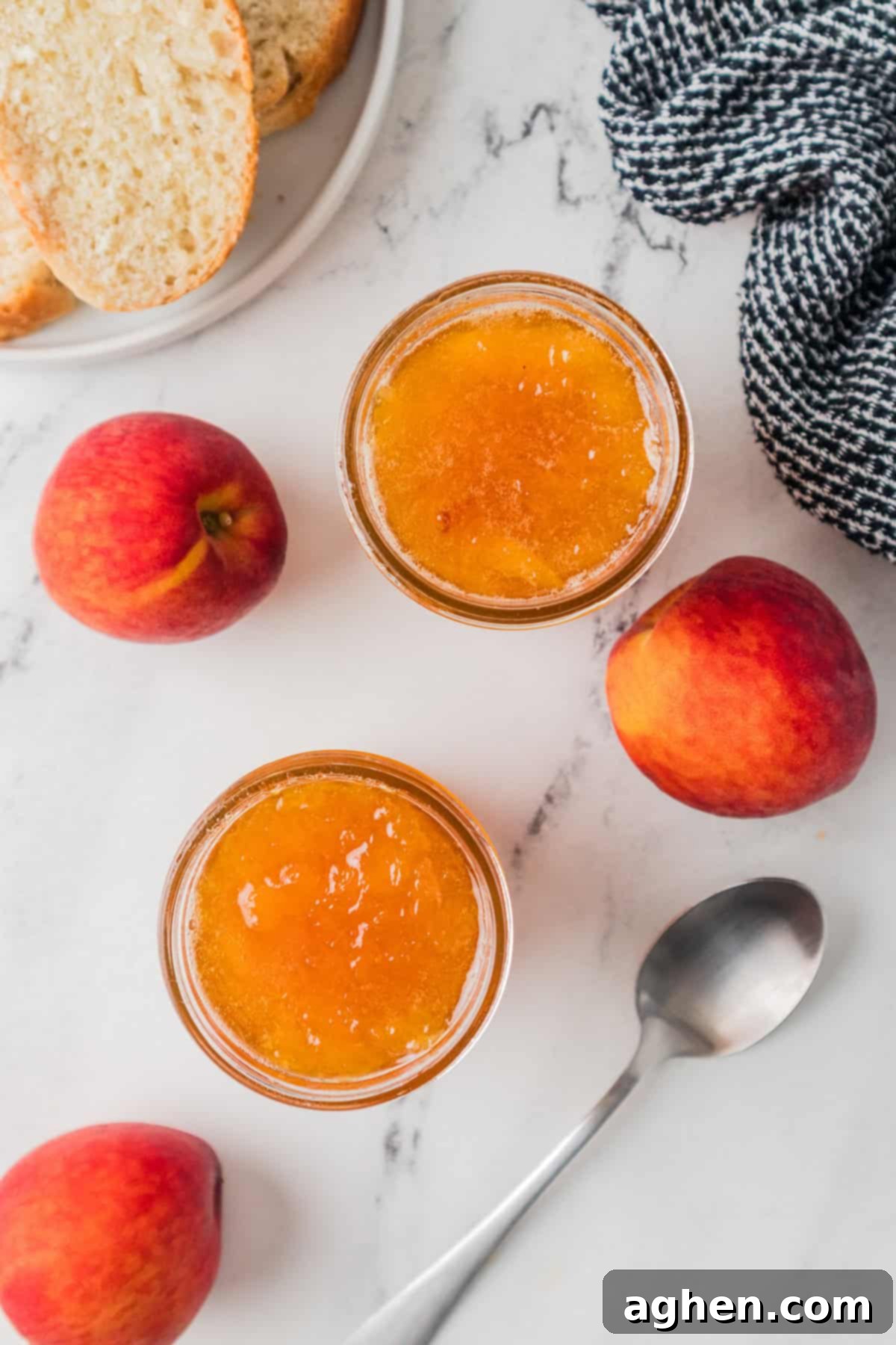 Overhead view showing two open jars of peach jam on a marble surface, surrounded by whole fresh peaches, a silver spoon, a black-and-white cloth, and a plate with slices of bread. Delicious homemade peach jam for breakfast.