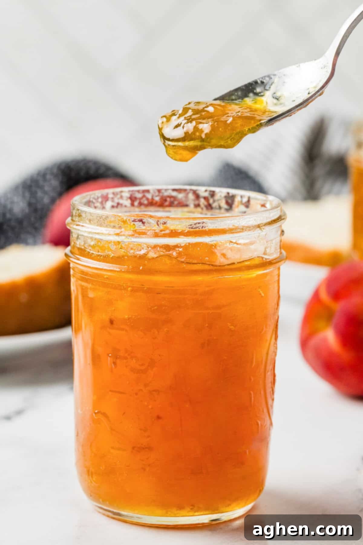Close-up of a spoon lifting thick peach jam from an open mason jar, with a peach and bread in the background. Ready to serve homemade peach jam.