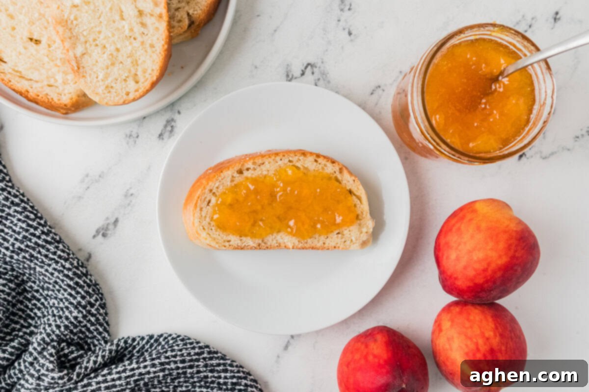 Slice of bread with peach jam on a plate next to a jar of homemade peach jam and peaches. Homemade peach jam on toast.