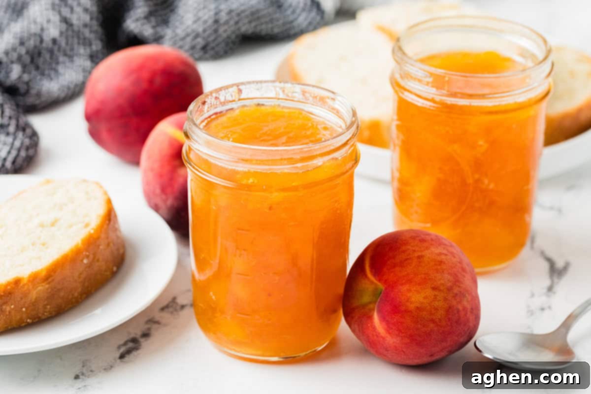 Two jars of peach jam placed on a marble surface with whole peaches, a spoon, and a plate with sliced bread in the background. Freshly made homemade peach jam ready to enjoy.