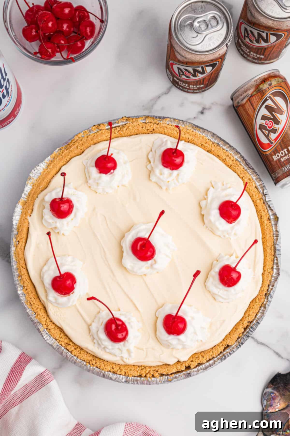 An overhead shot captures a full root beer float pie adorned with beautiful swirls of whipped cream and bright red cherries. It's surrounded by root beer cans and a bowl of maraschino cherries, setting the scene for serving.