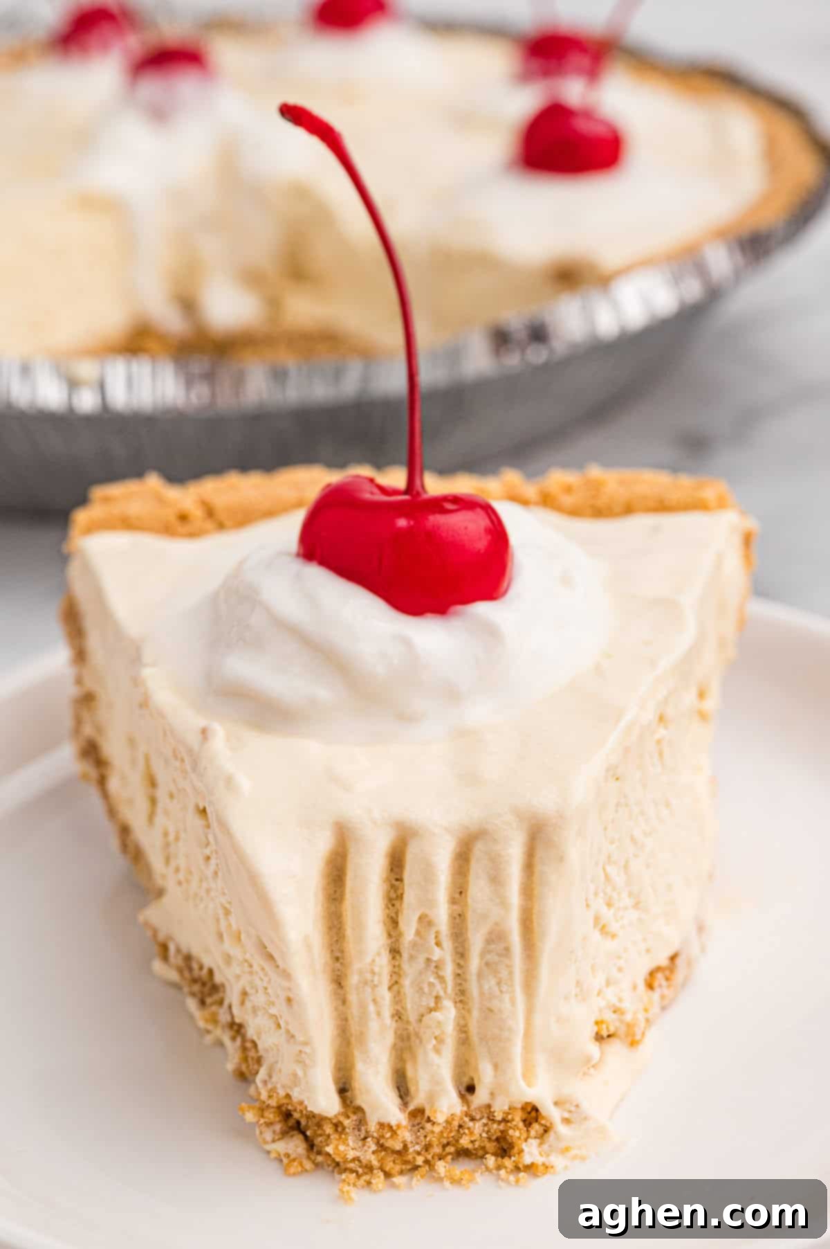 A close-up shot of a perfectly plated slice of root beer float pie. The creamy, light brown filling sits atop a golden graham cracker crust, finished with a generous dollop of whipped cream and a bright red maraschino cherry. Fork marks are visible in the slightly softened filling, inviting a bite.