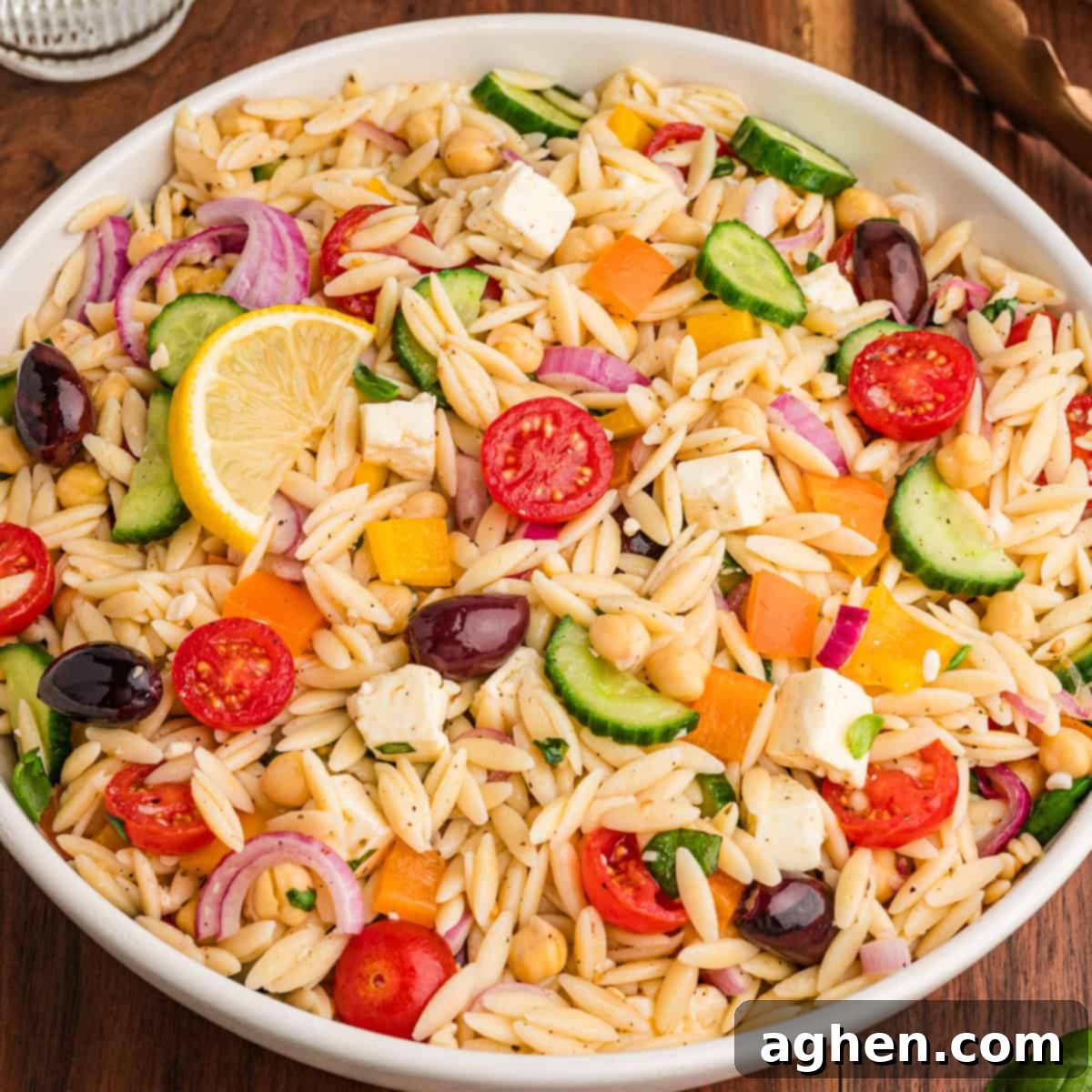 A bowl filled with orzo salad, highlighting visible ingredients such as halved cherry tomatoes, cucumber slices, red onion, chickpeas, yellow and orange bell pepper pieces, kalamata olives, cubed feta, and chopped herbs, with a lemon wedge on top.