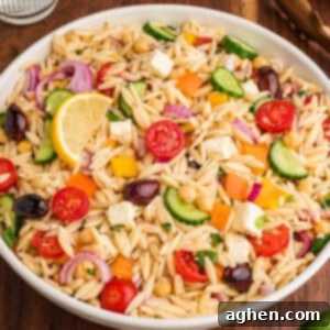 A bowl filled with orzo salad, highlighting visible ingredients such as halved cherry tomatoes, cucumber slices, red onion, chickpeas, yellow and orange bell pepper pieces, kalamata olives, cubed feta, and chopped herbs, with a lemon wedge on top.