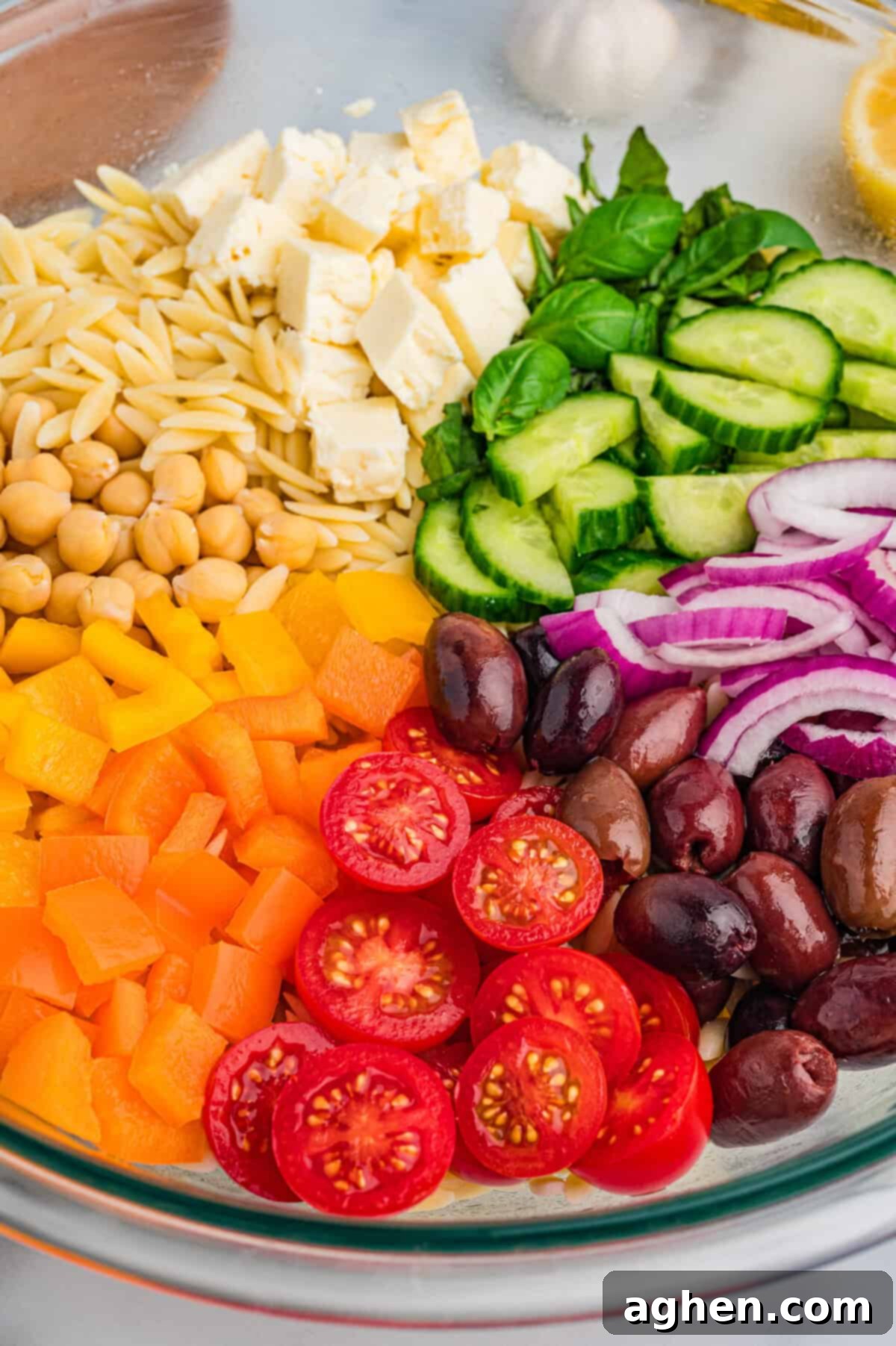 lose-up image of a glass mixing bowl with orzo salad ingredients placed in neat sections, including cherry tomatoes, kalamata olives, orange and yellow bell peppers, chickpeas, feta, basil, cucumber, and red onion.