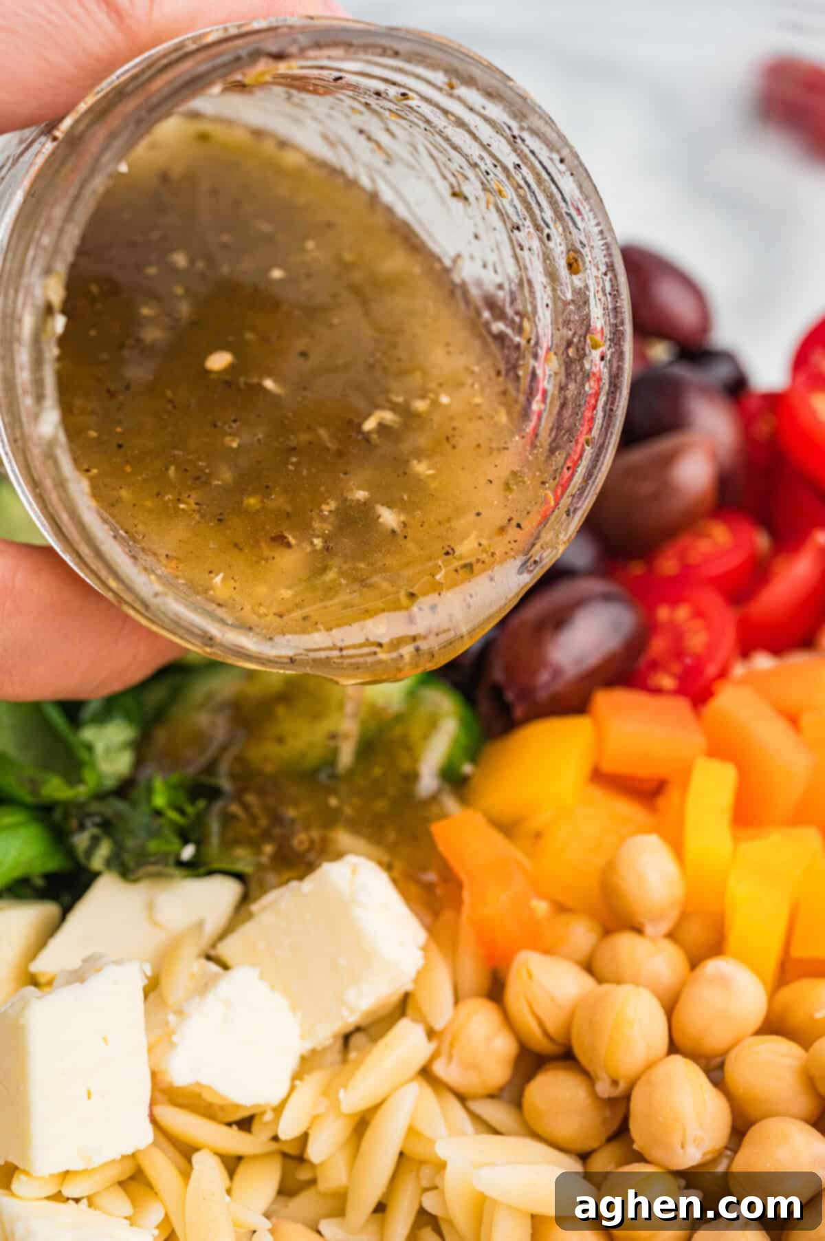 Salad dressing being poured from a jar over orzo salad ingredients.