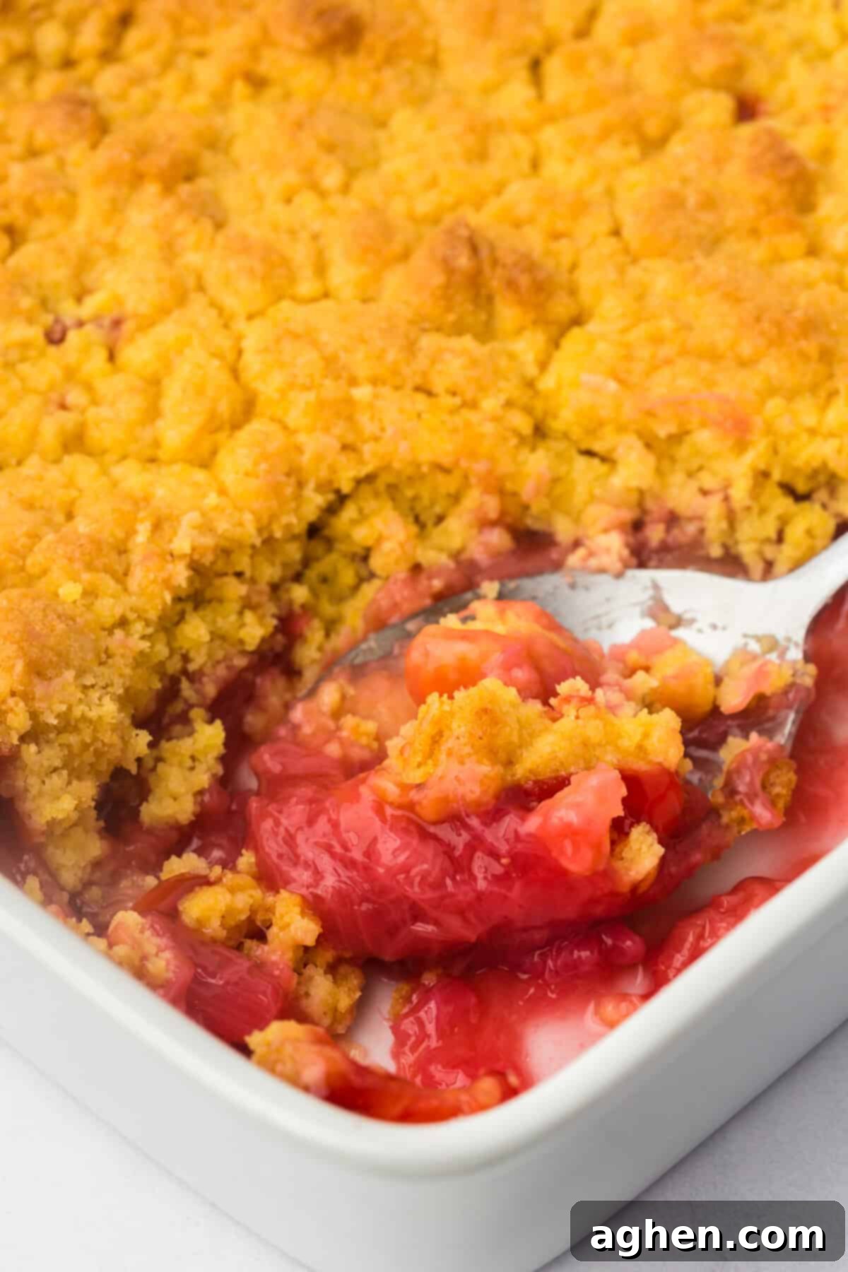 Easy Rhubarb Dump Cake 3 A spoon lifting a serving of strawberry rhubarb dump cake from the baking dish, showing the inviting golden crust and bright pink-red filling, ready to be served.