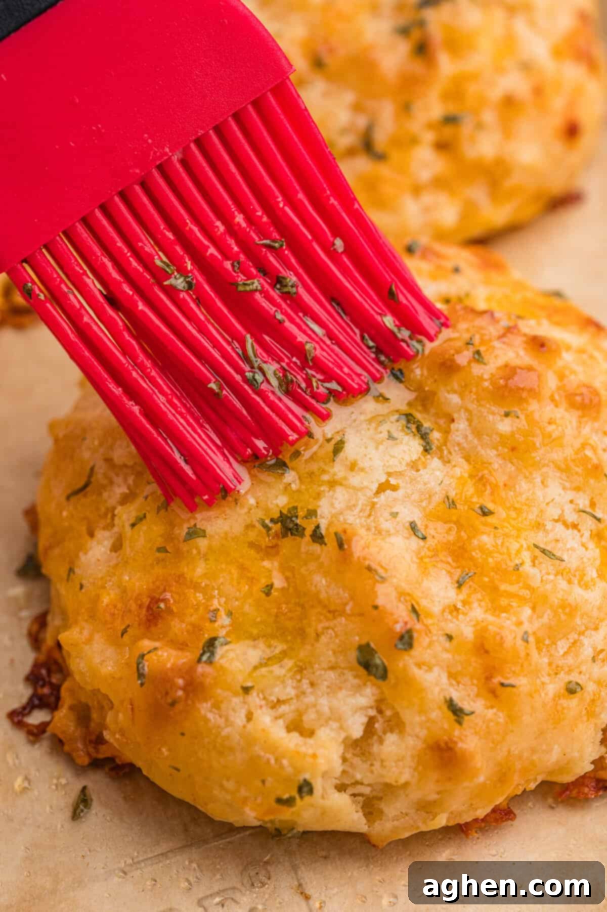 Fluffy Cheddar Biscuits 3 A red silicone brush applies melted butter with herbs onto the top of a freshly baked cheddar biscuit, enhancing its golden, textured surface.