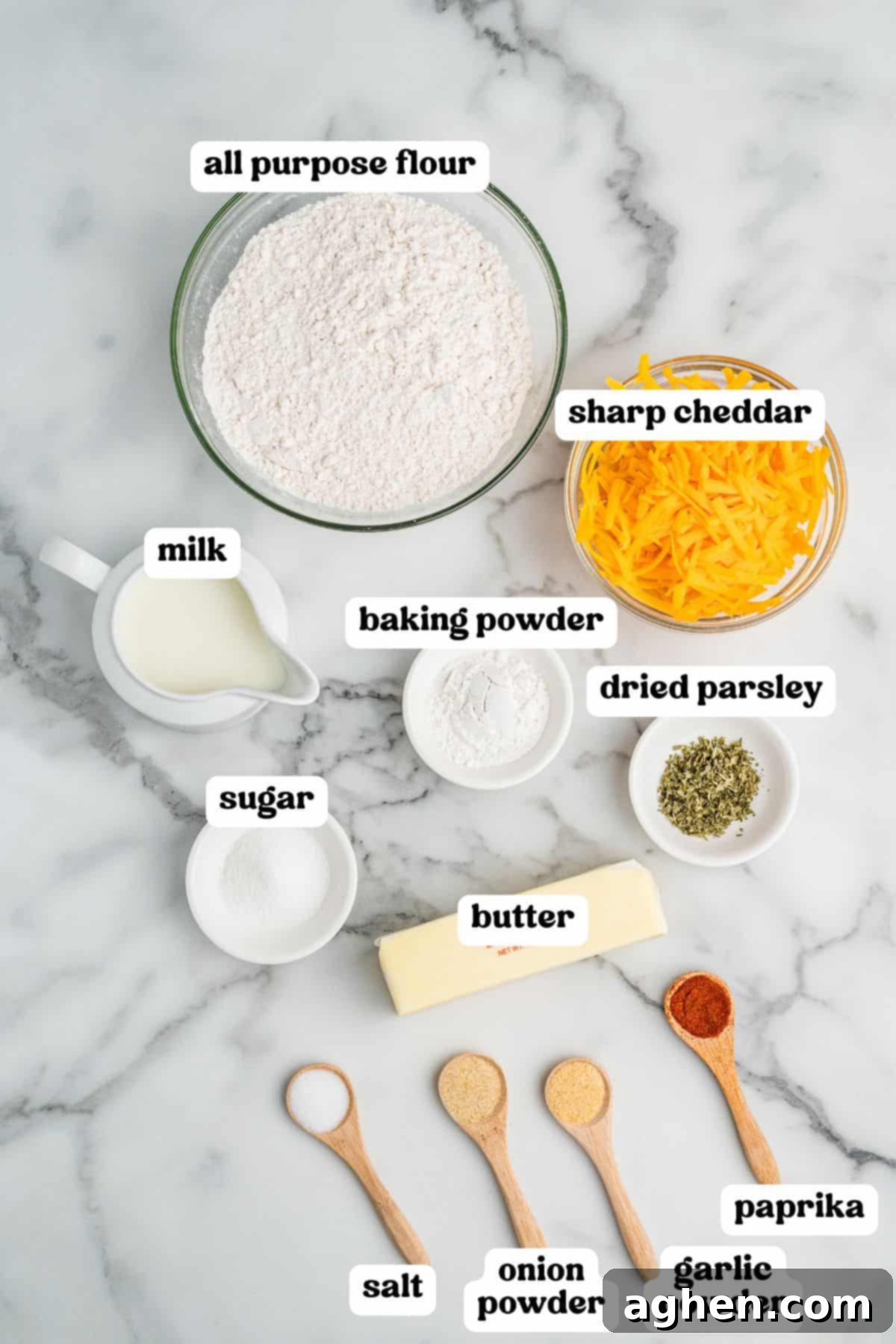 Fluffy Cheddar Biscuits 4 Overhead view of ingredients for cheddar biscuits arranged on a marble surface, each labeled: all-purpose flour, sharp cheddar, milk, baking powder, dried parsley, sugar, butter, salt, onion powder, garlic powder, and paprika.