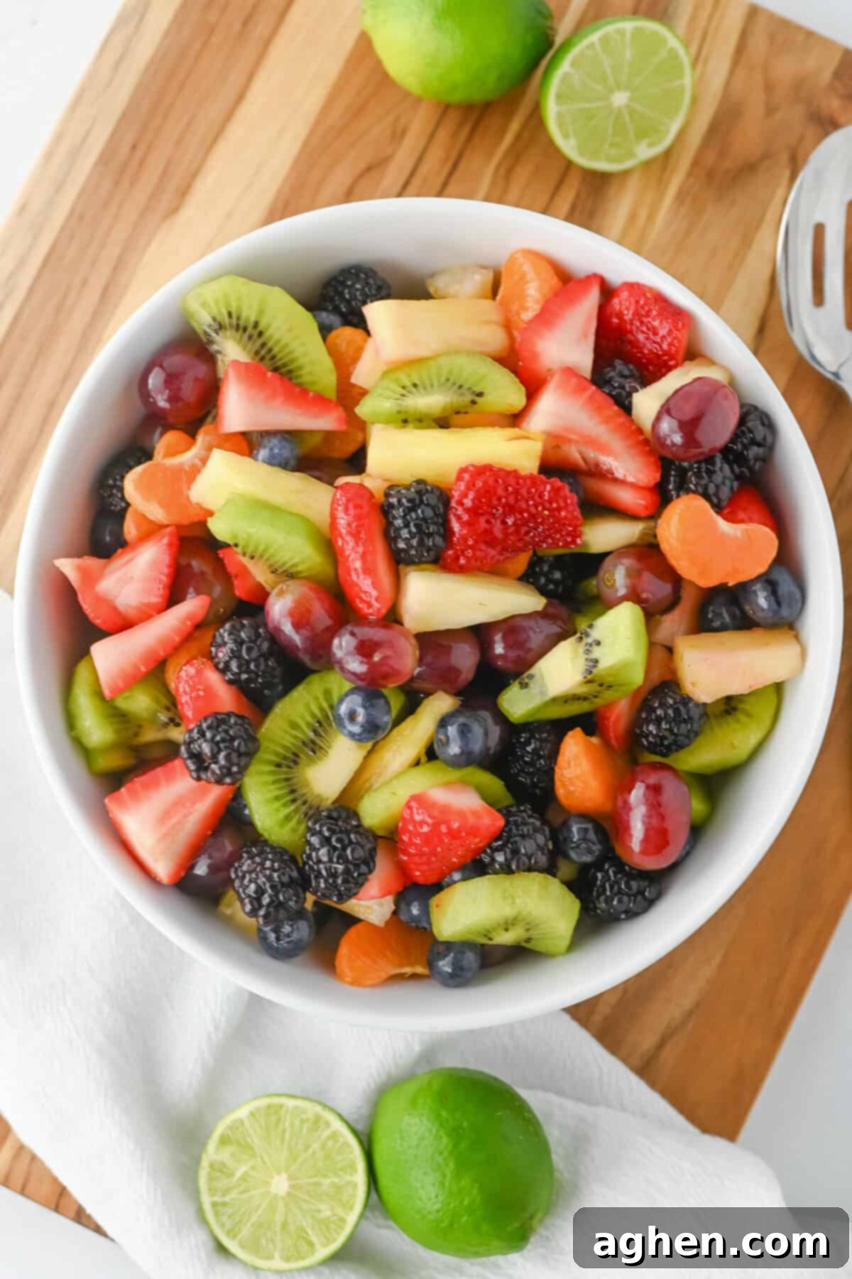 Fruit salad with honey lime dressing in a white bowl with sliced strawberries, kiwi, pineapple, grapes, blackberries, blueberries, and clementine slices. The bowl is set on a wooden board with halved limes nearby.