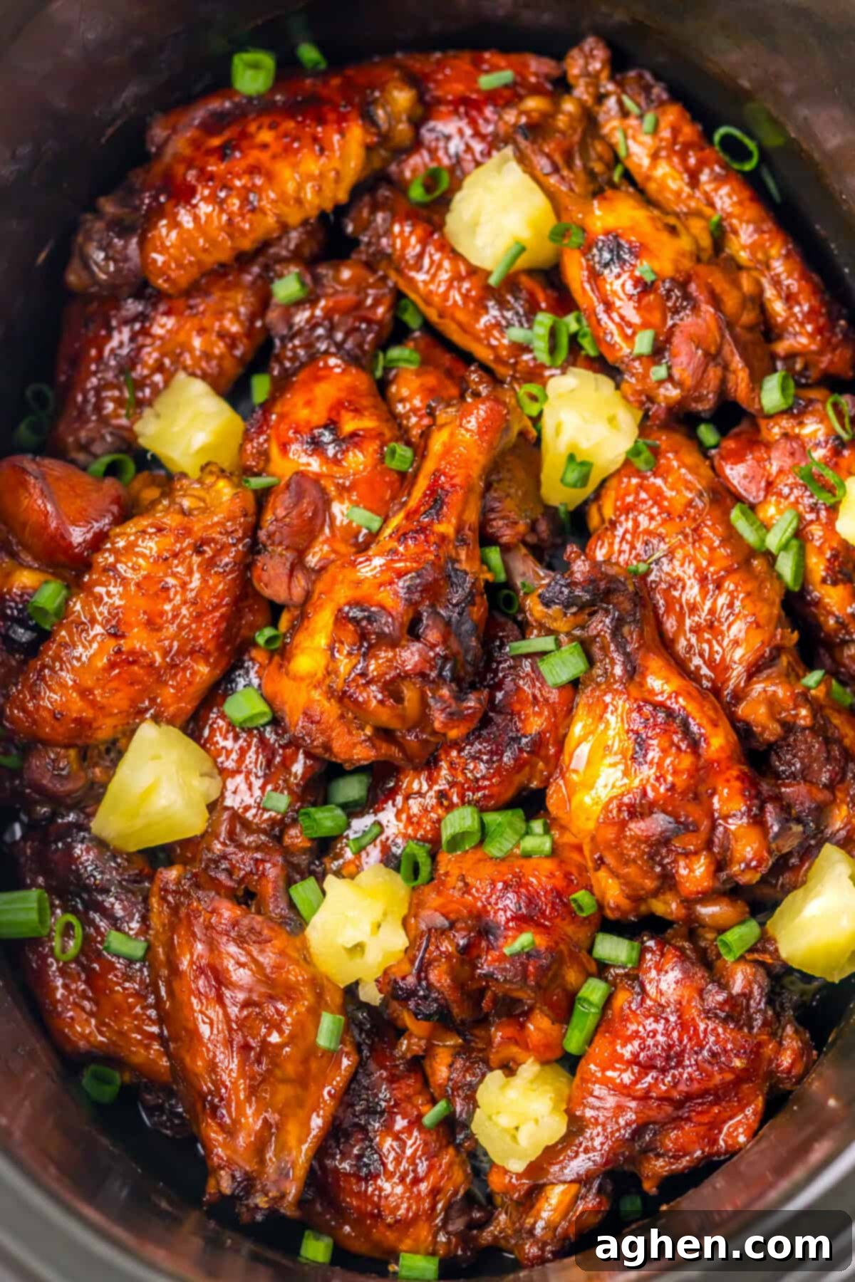 A crockpot packed with glazed Hawaiian wings. The chicken wings are topped with chunks of pineapple and chopped scallions.