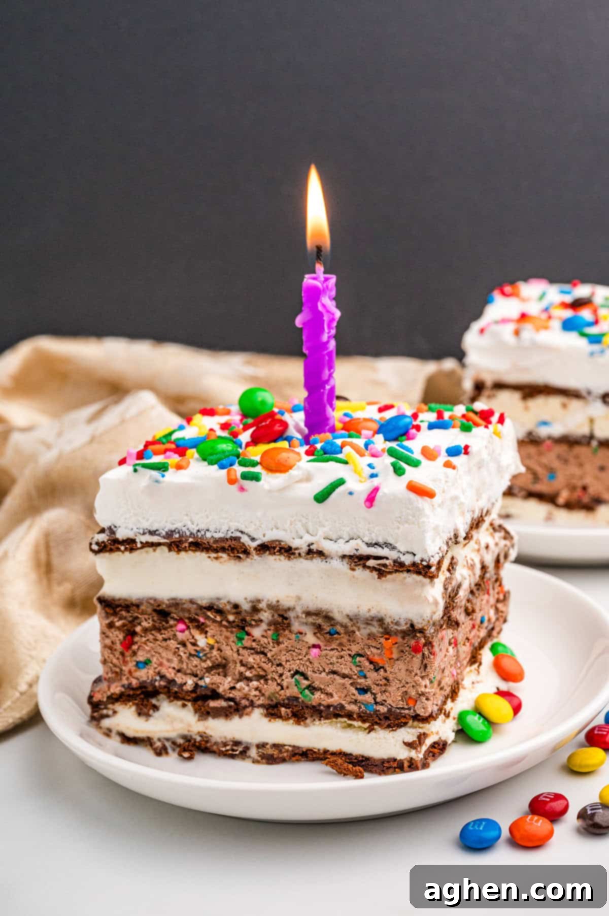 A vibrant slice of ice cream sandwich cake, topped with cool whip, colorful sprinkles, mini M&Ms, and a lit purple candle, ready for a celebration.