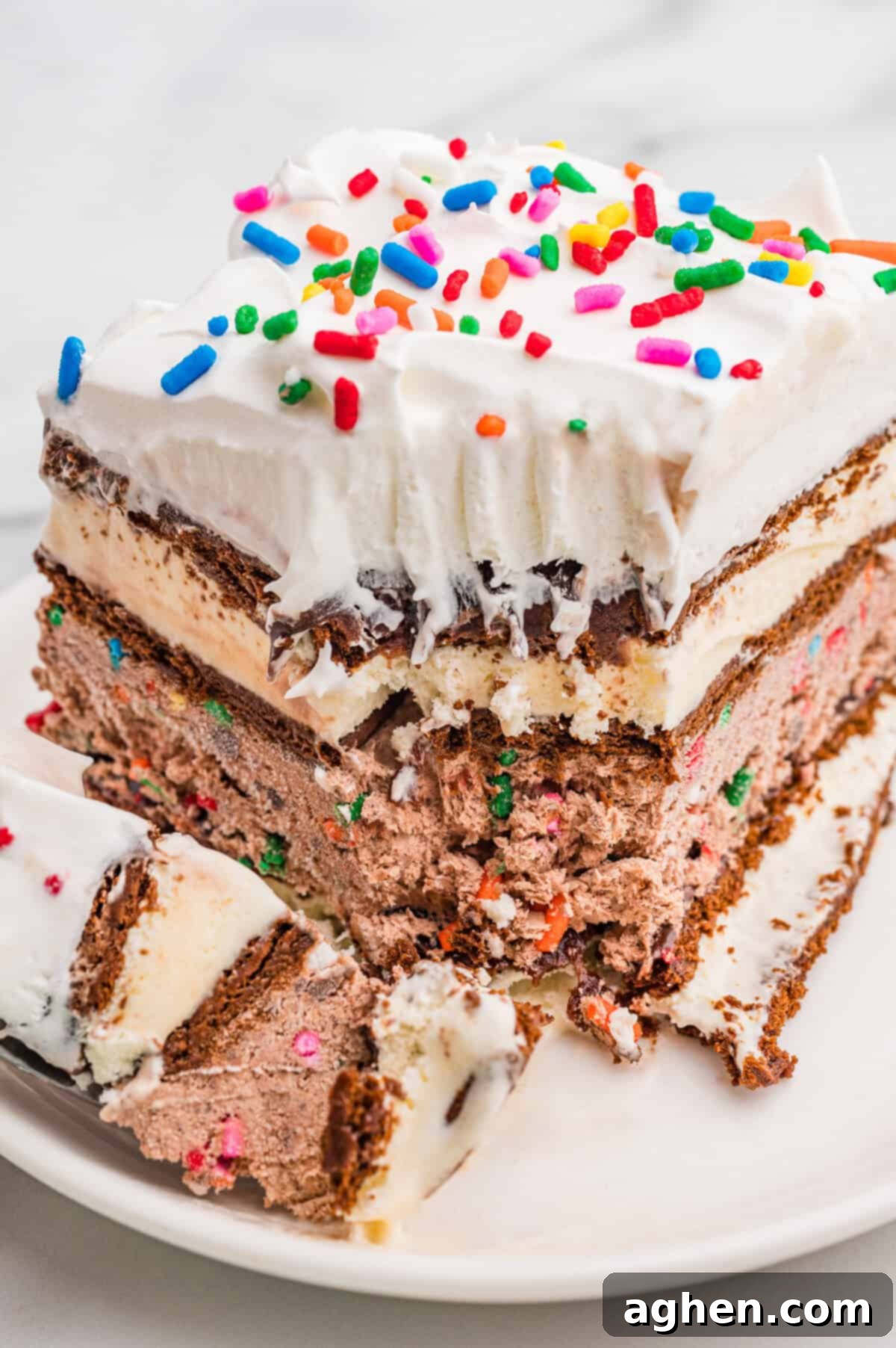 A close-up of a partially eaten slice of ice cream sandwich cake, showcasing the distinct layers of ice cream sandwiches, chocolate pudding, and whipped topping, all adorned with colorful sprinkles.