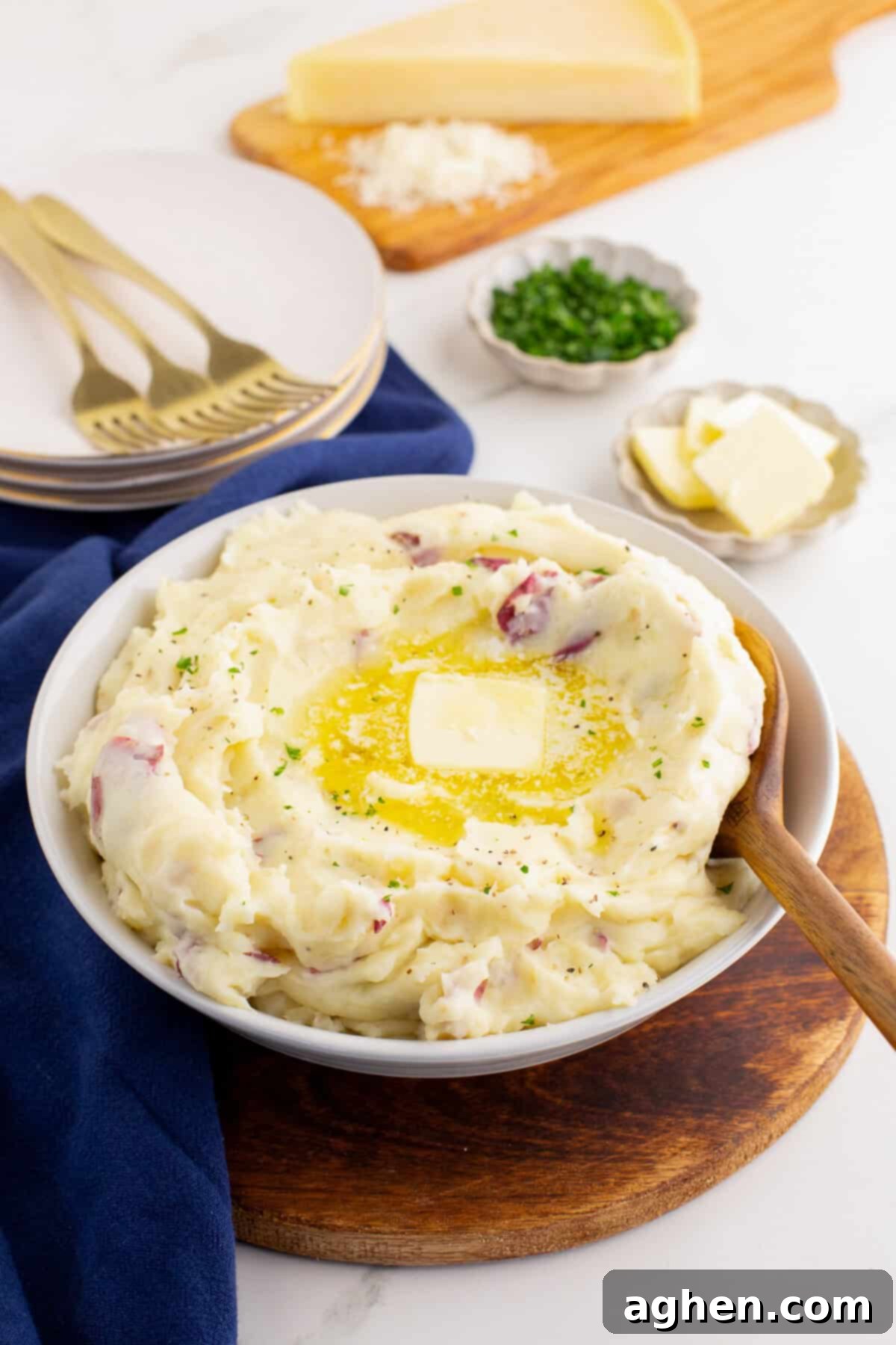 White bowl filled with red skin mashed potatoes, topped with a pat of melting butter and sprinkled with herbs, set on a wooden board with plates, forks, Parmesan cheese wedge, chopped chives, and extra butter in the background.