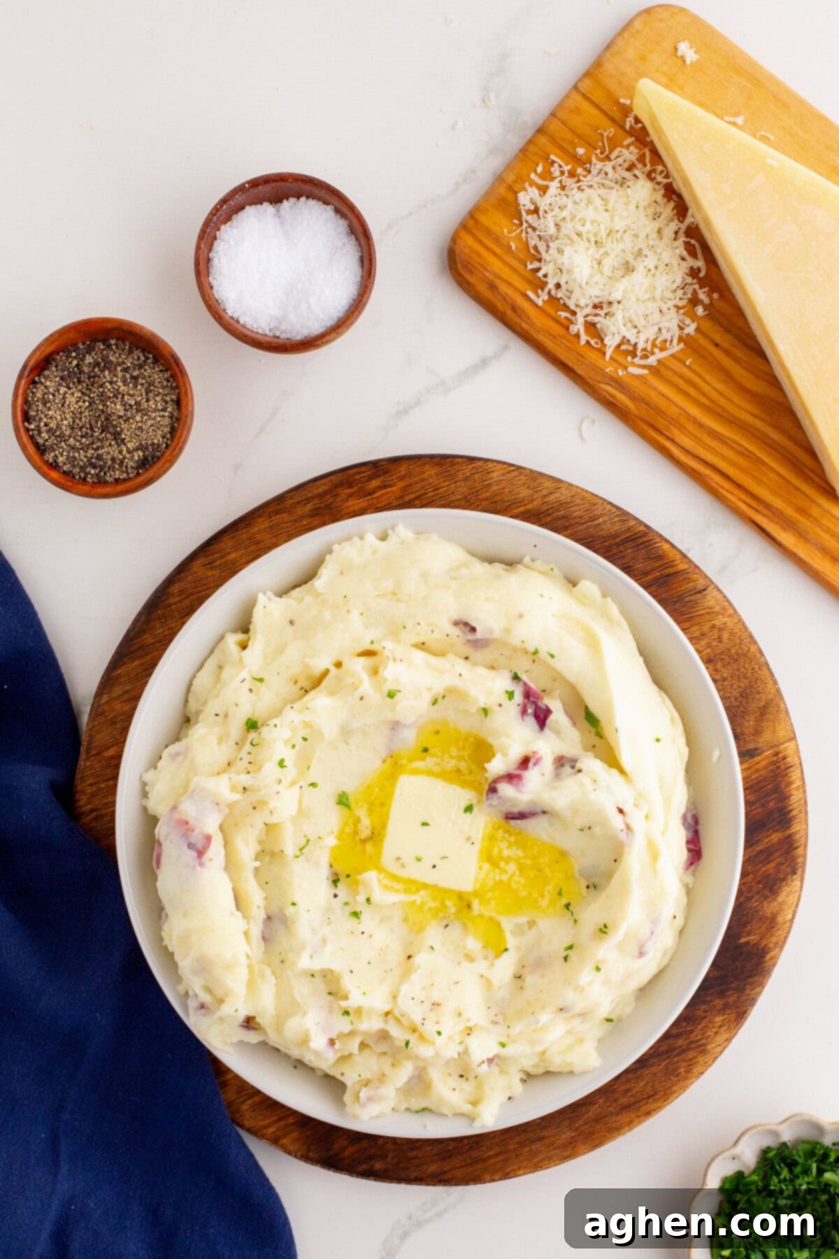 Overhead photo of a white bowl of mashed potatoes with red skin potatoes, garnished with a pat of melting butter and herbs, set on a wooden board with bowls of salt and pepper, a wedge of Parmesan cheese, and shredded cheese on a wooden cutting board.