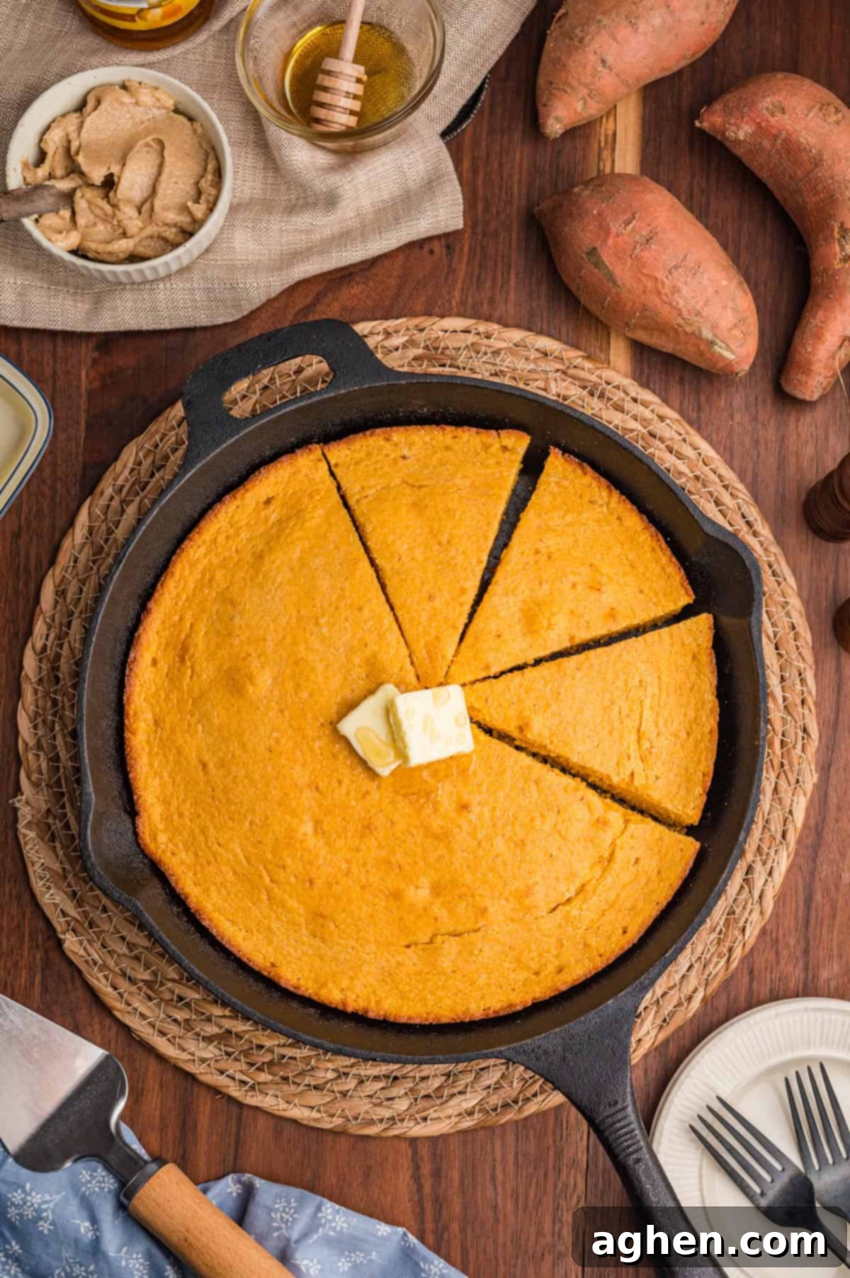 Overhead view of a cast iron skillet filled with sweet potato cornbread, sliced into wedges. A pat of butter is melting in the center, surrounded by fresh sweet potatoes, a honey jar, cinnamon butter, and a serving spatula on a rustic wooden table.