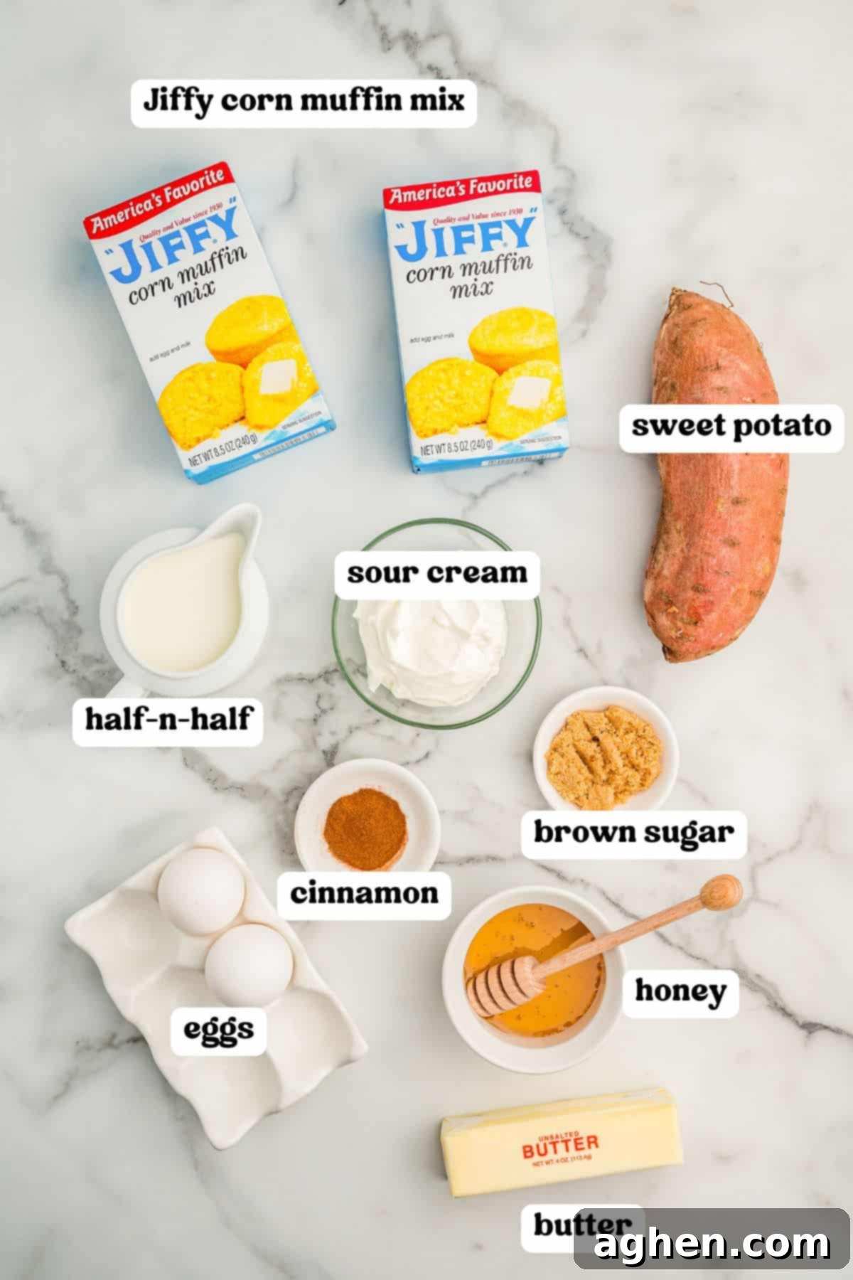 Labeled ingredients for sweet potato cornbread including two boxes of Jiffy corn muffin mix, a sweet potato, sour cream, half-and-half, cinnamon, brown sugar, honey with a dipper, butter, and two eggs on a white background.