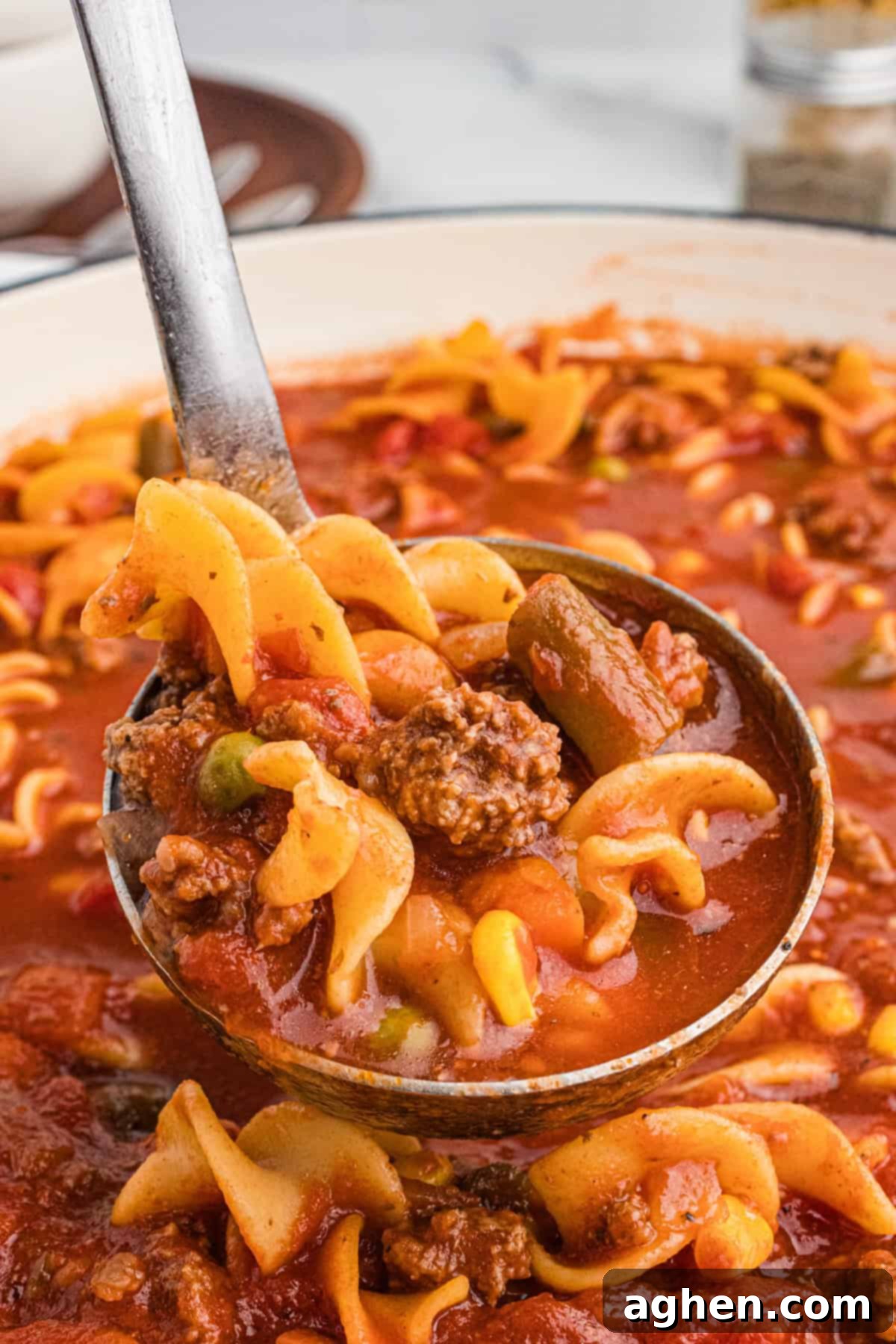 Close-up of a ladle full of hamburger noodle soup showing egg noodles, ground beef chunks, corn, carrots, green beans, peas, and tomatoes in a red broth.