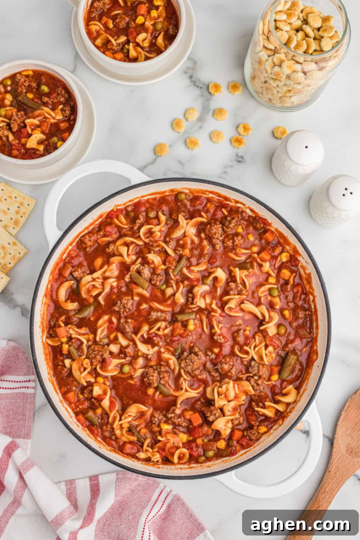 Top-down image of a white pot filled with hamburger noodle soup, featuring ground beef, egg noodles, mixed vegetables, and tomato-based broth. Two smaller bowls with soup are nearby, along with a glass jar of oyster crackers, scattered crackers, salt and pepper shakers, and saltine crackers.