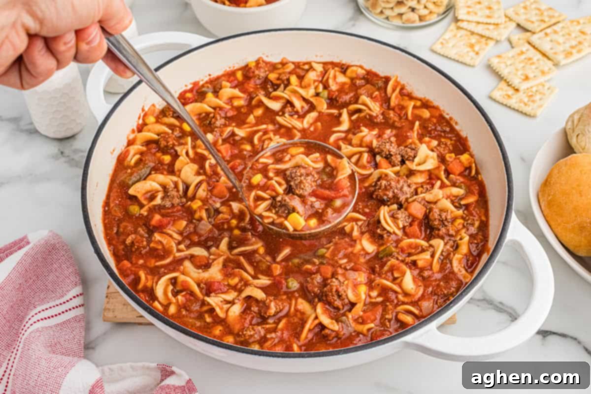 A ladle scooping hamburger soup from a large pot surrounded by crackers and rolls. The soup contains ground beef, egg noodles, corn, peas, green beans, carrots, and diced tomatoes.