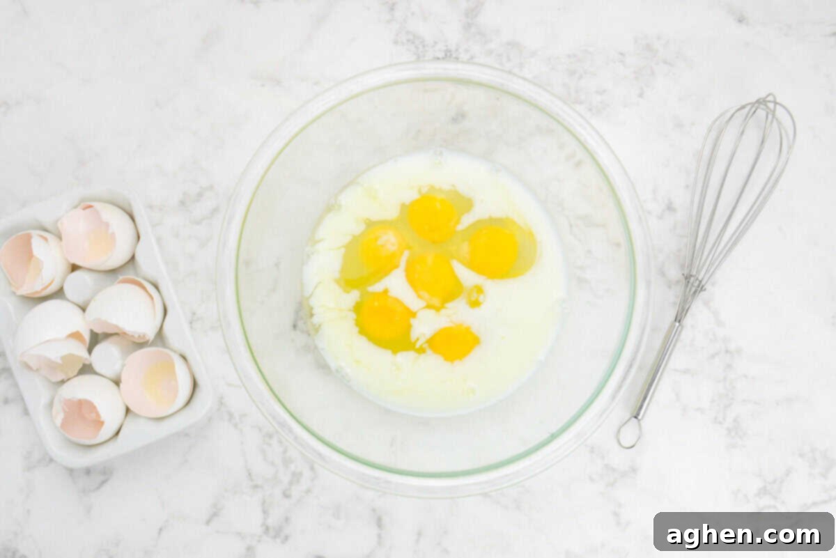Smart Start Breakfast Bake 5 A large, transparent mixing bowl contains uncooked eggs and milk, patiently waiting to be whisked. Adjacent to the bowl, a shiny metal whisk and the remnants of cracked eggshells hint at the initial steps of the recipe preparation.