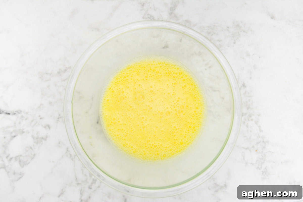 Smart Start Breakfast Bake 6 A large, clear mixing bowl is filled with a smoothly whisked mixture of eggs and milk, displaying a uniform, pale yellow color, indicating it's perfectly combined and ready for the next ingredients.