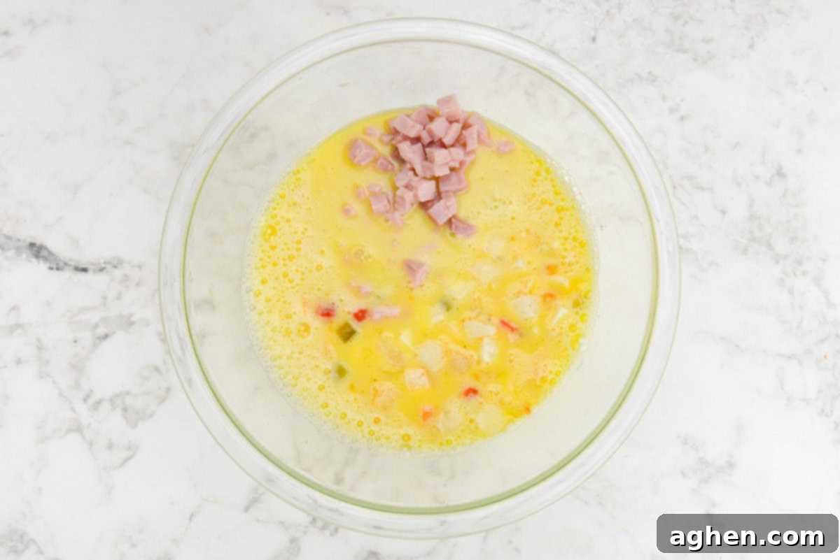 Smart Start Breakfast Bake 7 In a spacious mixing bowl, the whisked eggs are now joined by small, colorful pieces of bell peppers and potatoes that float near the surface. A mound of diced ham is distinctly piled in the center, not yet fully mixed into the creamy egg base.